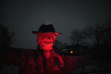 Terrible scarecrow in dark cloak and dirty hat Illuminated with red light stands near the house at...