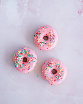Overhead view of three pink glazed donuts with rainbow sprinkles on a light pink background