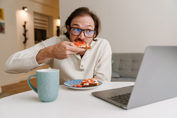 Ginger bristle man in eyeglasses working with laptop while having dinner