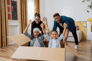 Parents have fun driving cute little son and beautiful daughter in cardboard box play in living room on relocation to new home family feel overjoyed involved in fun activities of moving in together