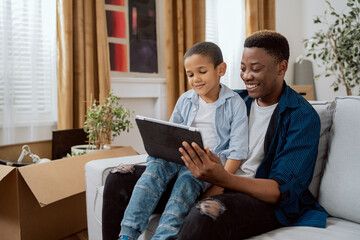 Fototapeta premium A proud father sits on the couch holding son on his lap, a tablet in hand, they take a selfie, a picture for social media, they smile