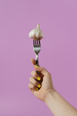 Female hand holding garlic on fork. Minimal composition with purple background. Creative healthy...