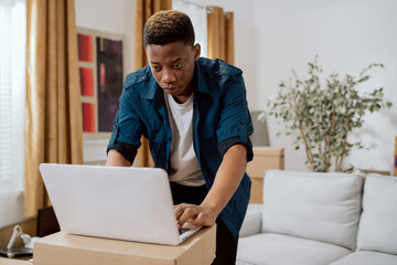 Man moves into new apartment, no furnishings except couch, laptop stands on cardboard boxes, man browses online furniture stores, looking for dresser, closet, decorations to buy