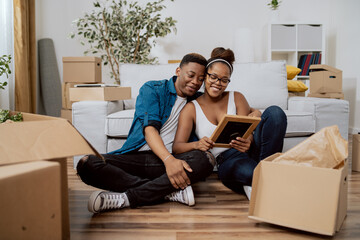 Married couple after moving to new house unpack boxes sitting on floor, woman shows husband found photo in frame from their wedding, man embraces wife, they smile reminisce