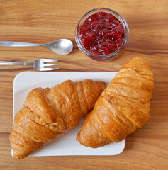 Two close-up croissants lie on a white plate next to a jar of berry jam. The photo is square.