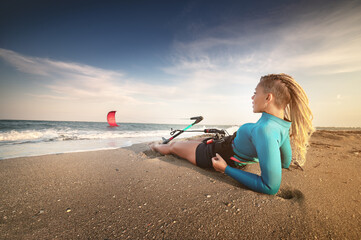 Attractive caucasian woman with dreadlocks on her head in a wetsuit lies on a sandy beach and holds her kite. Water sports. Kite surfer on vacation. Copy space