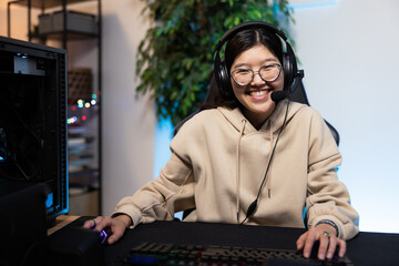 Joyful laughing professional Asian girl gamer wearing headphones with microphone playing online video games with friends colorful neon lights computer in living room at home.