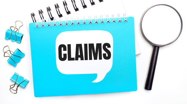 Office table with blue notepad, paper clips, magnifying glass and white card with text CLAIMS. Top view, flat lay. Stylish workplace