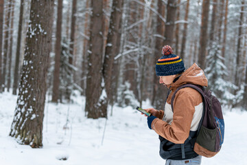 Fototapeta na wymiar Young man in knitted hat with backpack looking at map on smartphone while hiking in winter forest using modern technology navigation
