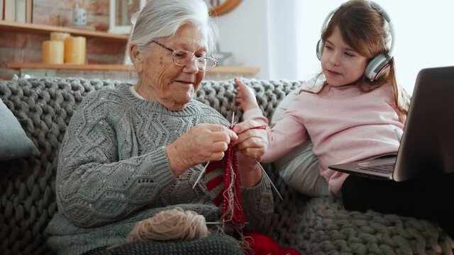 Old Grandma knits and her granddaughter uses a laptop to surf the Internet while listening to music