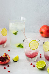 Refreshing pomegranate drink in glasses with pomegranate seeds and ice, lime and glass straws, jug of cold water with citrus, cut pomegranates with seeds