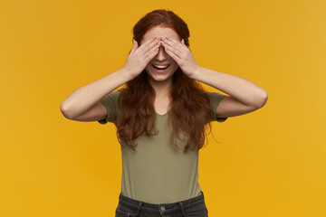 Indoor portrait of young ginger female joyfully close her eyes with palms, smiles broadly and feels happy. isolated over yellow background