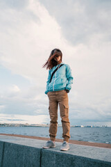 A young woman in windy weather stands against the background of the city embankment. © Alex Photo