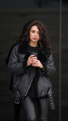 Girl in black clothes with a cup of coffee. The girl is standing next to the glass wall. Curly hair. Stylish clothes jacket, leggings.