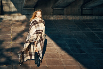 Full length portrait of pretty young woman wrapped in blanket posing ib sun lights over urban...