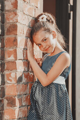 a young brunette girl in a blue polka dot dress stands against the wall in the summer