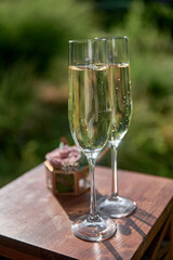 Sparkling wedding glasses with champagne and wedding rings in box in blur on wood table outdoors, copy space