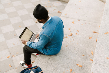 Black man with headphones working with laptop while sitting on bench