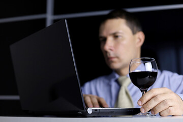 Man with glass of red wine in hand, sitting at laptop.  Remote work at home or relax at office with alcohol drink