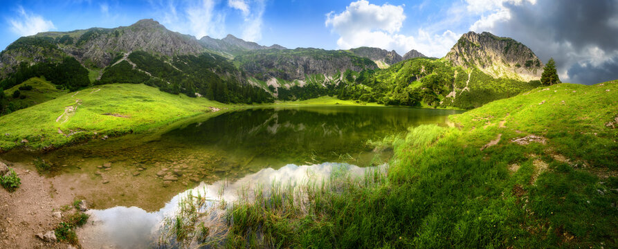 Mountain lake panorama with green meadows, blue sky, dramatic clouds and reflections on the clear water in the Alps