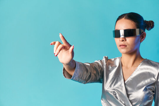 Trendy ethnic woman in futuristic VR goggles touching imaginary screen in studio
