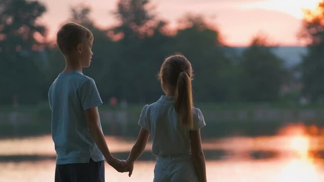 Happy siblings standing together holding hands in summer park. Young children brother and sister relaxing outdoors. Family love and relationship concept