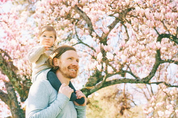 Happy young father carrying child on his shoulders, toddler girl with dad in spring blooming park...