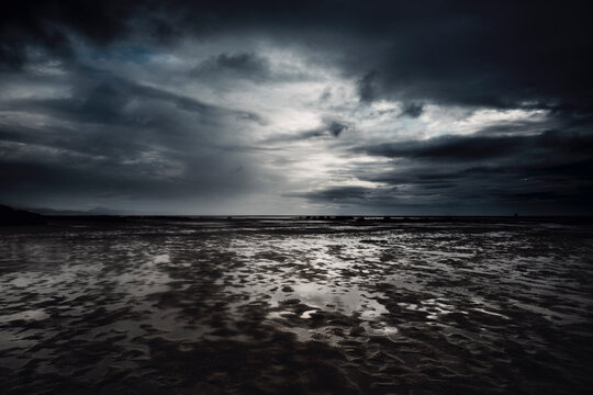 Dramatic cloudscape and flood beach in dark night weather. Stormy and rainy sky with ocean in background. Epic seascape with white sunset light and black clouds.