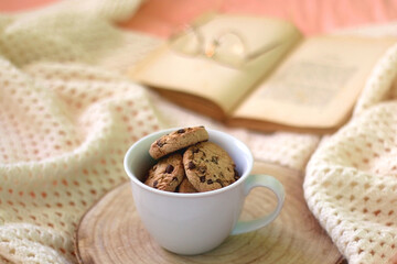 Fototapeta na wymiar Mug filled with chocolate chip cookies, open book, reading glasses and knitted blanket on the bed. Selective focus.