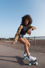Happy skater girl with afro hair skating at promenade. She is smiling, looking at camera and wearing spring clothes. © 1 MEDIA