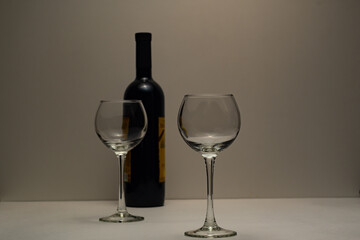 Two empty, transparent glasses and a bottle of good red wine. A bottle of red wine, two transparent wine glasses, on legs, on a light base, with a light background.