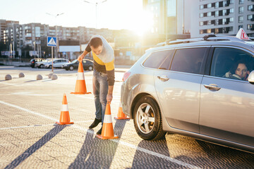 Male instructor teaching learner driver to park a car on the training ground with traffic cones at...