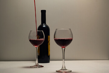 Two glasses filled with red wine, a drink is poured into one of the glasses. A bottle of red wine, one filled glass, the second glass is filled, on a light base, with a light background.