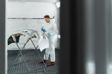 Car painter in protective clothes and mask painting and varnishing automobile bumper in paint spray booth