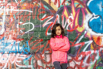Young girl with long dark hair in a pink jacket standing in front of a graffiti wall.