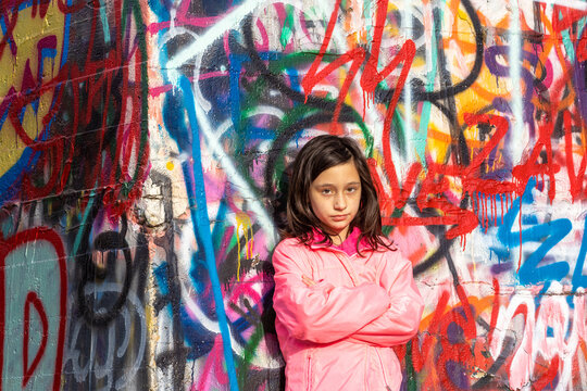 Moody pensive with deep emotion young girl with long dark hair in a pink jacket standing in front of a bright colorful graffiti wall.