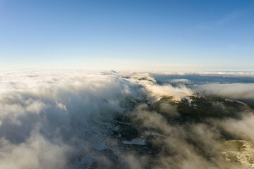 Aerial view from above of white puffy clouds covering snowy mountain tops in bright sunny day