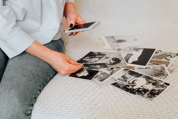 Nostalgia and memories concept. Close-up of woman taking photos on a smartphone camera of old black-and-white photographs from family album while sitting on sofa, indoors. Selective focus