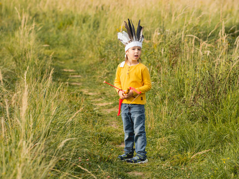 Little Boy Is Playing American Indian On Field. Kid Has Handmade Headdress Made Of Feathers And Bow With Arrows. Costume Role Play. Outdoor Leisure Activity. Fall Season.