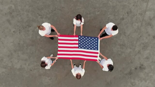 Team of boy and girl friends in jeans and white t-shirts unfolds bright-colored USA flag standing in circle on ground aerial view