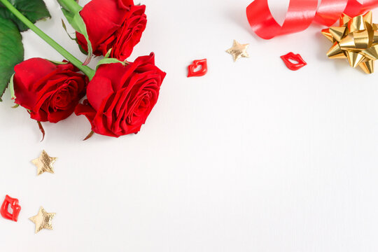 Festive background with a bouquet of red roses, gifts and gold confetti in the form of stars on a white wooden table top view. Empty template with copy space.