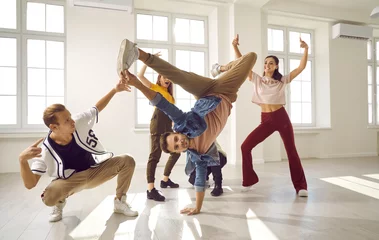 Fotobehang Flexible and energetic male dancer has fun with his dance group doing elements of breakdance movements. Young people in stylish youth casual clothes dance in bright dance hall with windows. © Studio Romantic