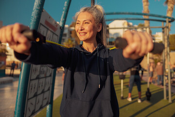 Obraz premium Portrait of sporty woman training with rubber bands at outdoor sportsground, smiling looking to the side