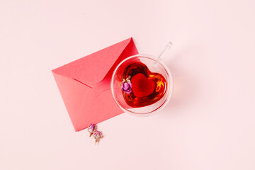 Natural herbal tea with rosebuds in a glass cup in the shape of a heart on a pink background. Black tea with a red envelope for Valentine's Day with space for text