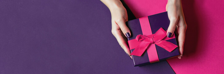 Women's hands with beautiful manicure hold a gift. Banner with a pink bow on a purple background....