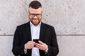 Positive urban business professional man in stylish suit using smartphone while standing outside