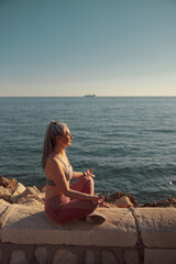 Fototapeta na wymiar Female in sports clothes sitting by the beautiful seashore in lotus position and training on sunny day