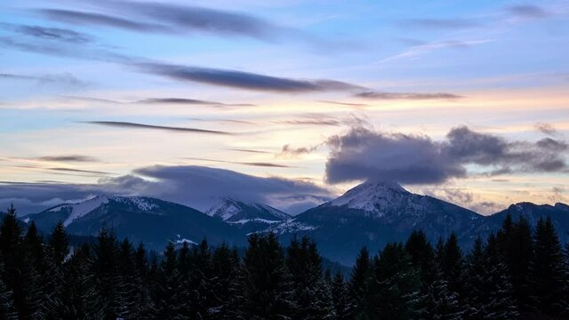 Clouds forming over the mountains at dusk. Trees in the foreground covered with snow. Colorful sky