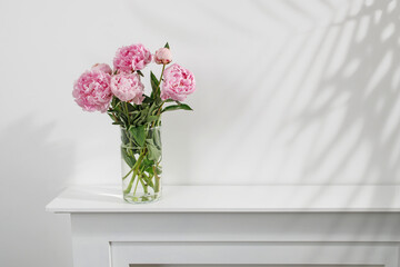 Red, pink peonies in a white vase on a table against a white wall background. Copy space