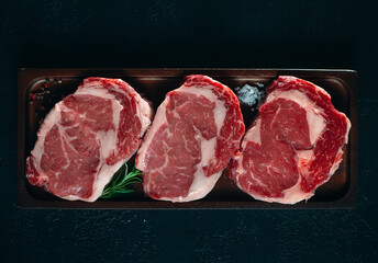 Three fresh raw rib eye steaks on a wooden board on a black background with salt, pepper and rosemary in a rustic style.